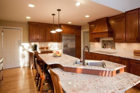 Remodeled kitchen with large uniquely shaped island. Woodgrain cabinets, white stone countertop, lowered section of island with regular dining chairs for seating.