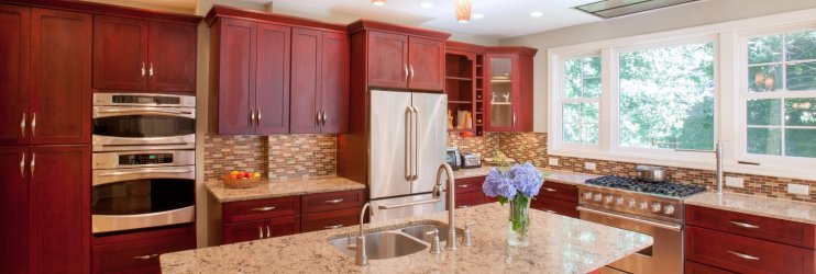Kitchen with cherry wood cabinets and tri-tone backsplash. Large island with double sink, large white double-hung windows to the right, above the stove.