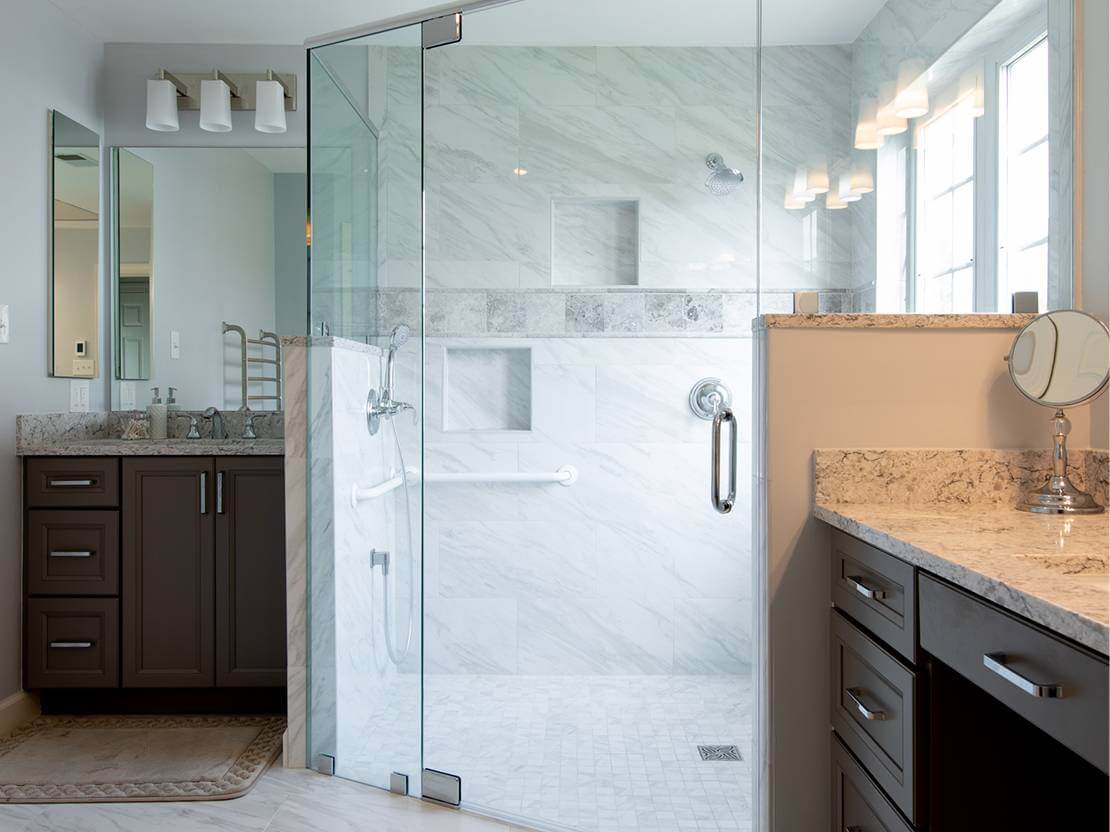 Close up on a glass door shower with granite tiling on the floor near two sinks.