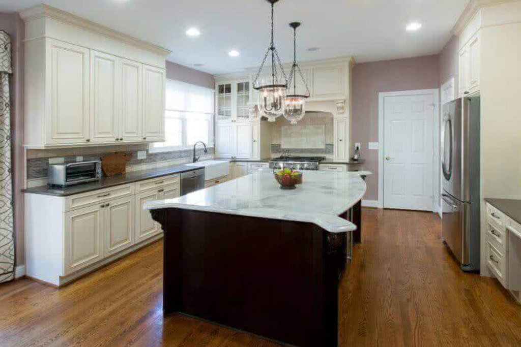 A large kitchen with an island in the middle. Wood floors, white cabinets around walls, island is black with white marble countertop and pendant lights above.