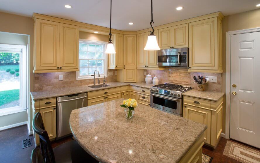 Remodeled kitchen with pendant and recessed lighting, centered kitchen island, and stainless steel appliances. Pale yellow cabinets. Tan quartz counters.