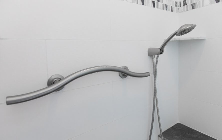 A curved grab bar on the wall of a shower, next to the detachable showerhead. White tile walls.