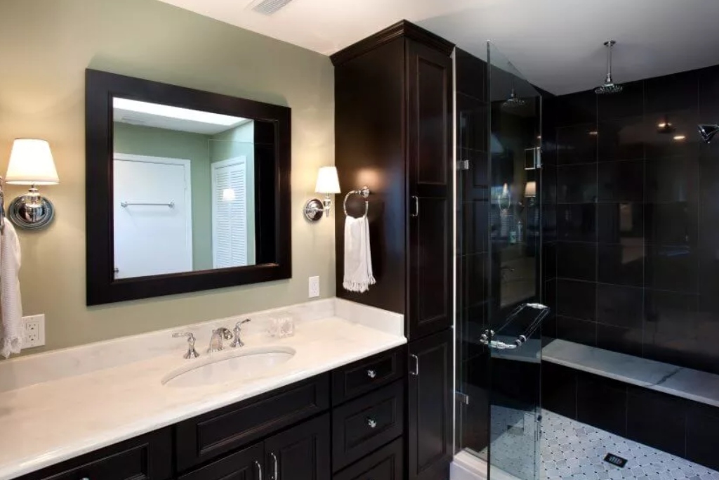 A bathroom with a sink, mirror, and shower stall. Black cabinets and black tile around shower. White countertop.
