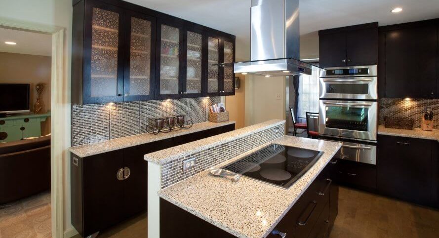 Custom kitchen remodel with dark cabinetry and light stone counter tops. Island with range. Glass tile backsplash. Glass upper cabinets.