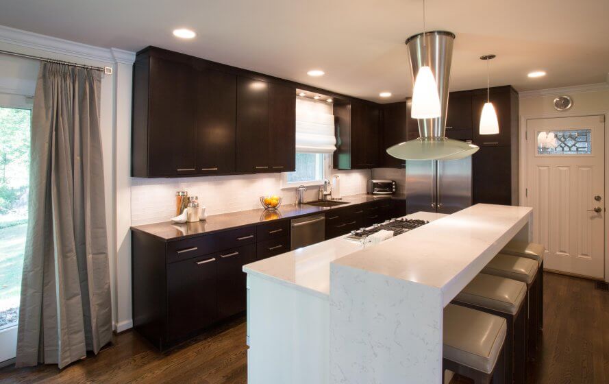 Remodeled kitchen with recessed and pendant lighting over a long white island with white stone waterfall countertop, and dark woodgrain modern cabinets along back wall.