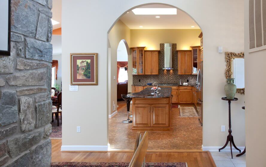 Remodeled kitchen viewed through arched doorway. Brown tile floors, woodgrain cabinets, dark stone countertops, and large island with seating in the middle.