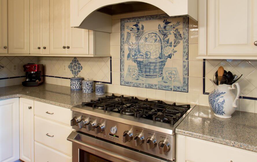French tile backsplash above stainless steel gas stovetop and oven. White cabinets, gray stone countertops.