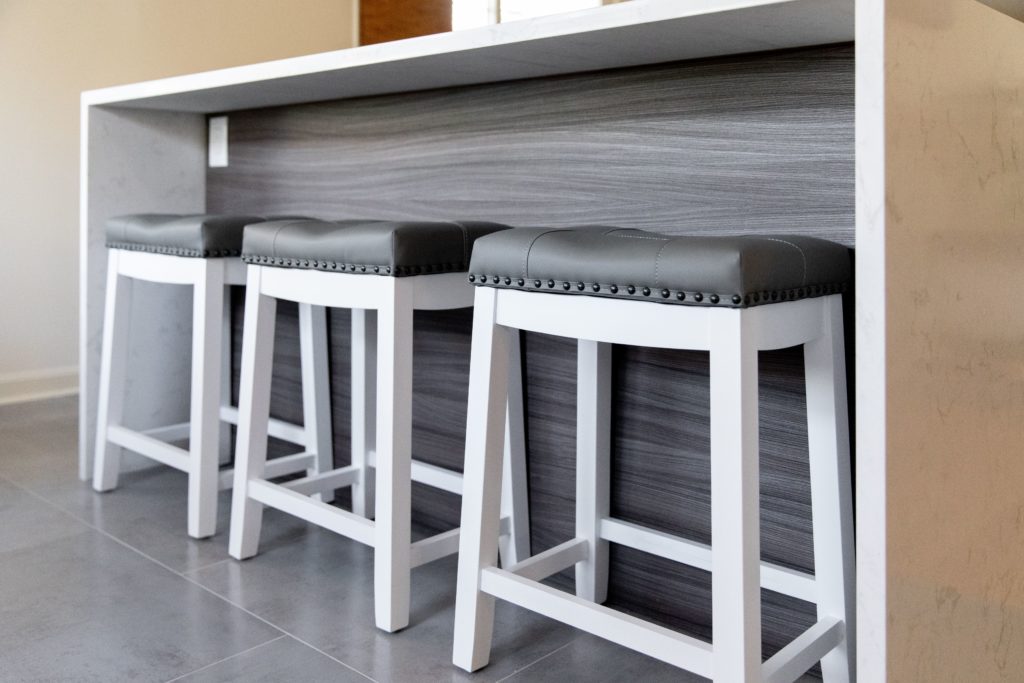 Island seating in a remodeled kitchen. White stools with gray cushioned seats, pushed under edge of an island with a white stone waterfall countertop.